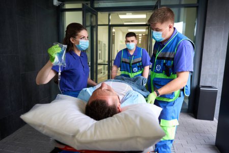 Foto de El hombre está siendo llevado a la sala de emergencias en una camilla especial, junto a él hay una enfermera sosteniendo un gotero - Imagen libre de derechos