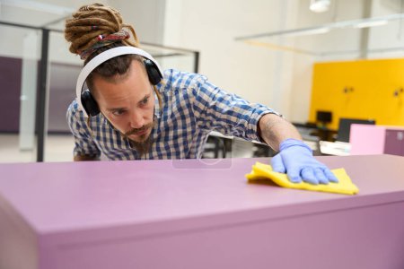Foto de El hombre que usa auriculares frota un casillero púrpura con un paño amarillo, usa un aerosol limpiador - Imagen libre de derechos