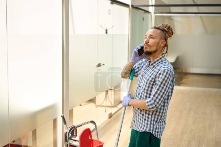 Photo for Informal guy is talking onphone in office corridor, next to his work gadgets are a red bucket and a mop - Royalty Free Image