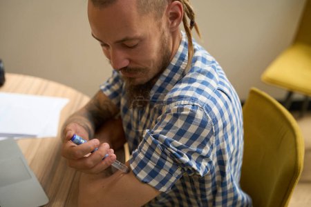 Photo for Guy with dreadlocks gives himself an injection in his arm, he sits at his desk - Royalty Free Image