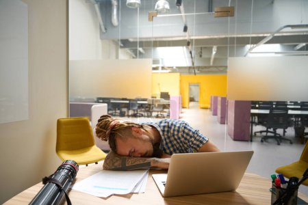 Foto de Freelancer en una camisa a cuadros sentado en su escritorio se quedó dormido mientras trabajaba debido a la mala salud - Imagen libre de derechos