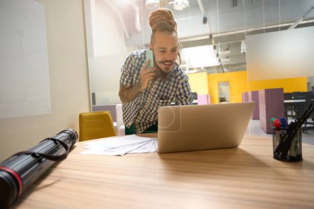 Photo for Guy with dreadlocks uses a phone and laptop to communicate, a freelancer is in a room with a modern interior - Royalty Free Image