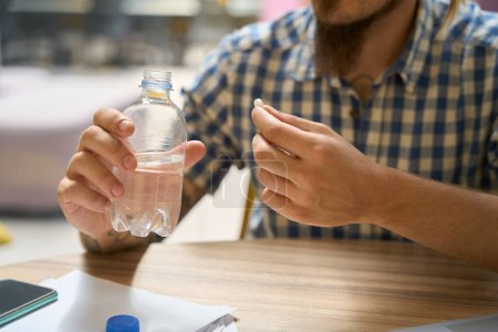 Photo for Guy in a plaid shirt holds a pill and a bottle of water in his hands - Royalty Free Image