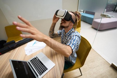 Téléchargez les photos : Homme informel utilise un casque de réalité virtuelle, il est assis à son bureau dans un espace de coworking - en image libre de droit