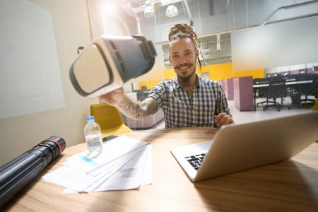 Foto de El joven sostiene los auriculares de realidad virtual en sus manos, está sentado en su escritorio en un espacio de coworking - Imagen libre de derechos