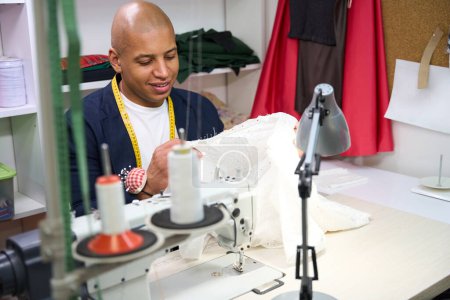 Foto de Retrato de cintura hacia arriba del sastre feliz sentado en la máquina de coser mirando la prenda en sus manos - Imagen libre de derechos
