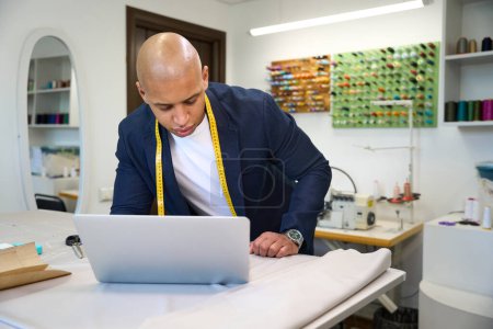 Téléchargez les photos : Concentré jeune tailleur se penchant sur son ordinateur portable sur la table de coupe dans l'atelier de couture - en image libre de droit