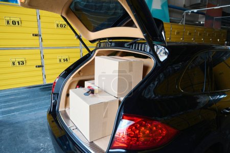 Photo for Shiny black car with an open trunk in a warehouse, there are many cardboard boxes in the trunk - Royalty Free Image