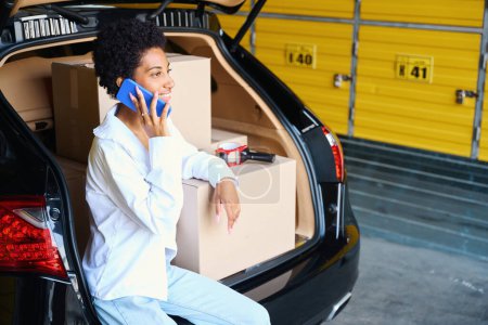 Téléchargez les photos : Femme afro-américaine parlant au téléphone, assise sur le bord du coffre, il y a beaucoup de boîtes en carton dans la voiture - en image libre de droit
