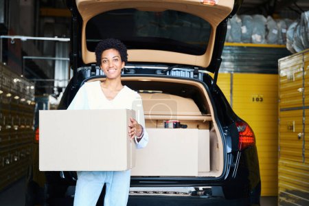 Téléchargez les photos : Femme en vêtements décontractés se tient dans un entrepôt avec une boîte en carton près de sa voiture - en image libre de droit
