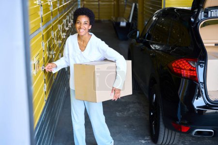 Téléchargez les photos : Jeune femme avec une boîte en carton ouvre une unité de stockage, à côté de sa voiture - en image libre de droit
