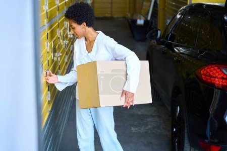 Téléchargez les photos : Jeune femme aux cheveux bouclés charge une boîte en carton dans un casier de rangement, à côté de sa voiture - en image libre de droit