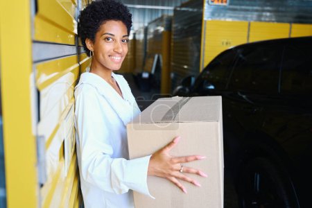 Téléchargez les photos : Charmante femme afro-américaine avec boîte en carton dans un entrepôt, à côté de sa voiture - en image libre de droit