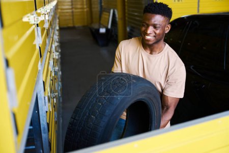 Téléchargez les photos : Pilote afro-américain charge des pneus de voiture dans un casier pour le stockage saisonnier, il utilise un service de stockage - en image libre de droit