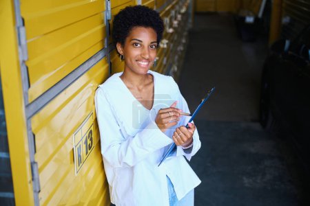 Téléchargez les photos : Femme afro-américaine se tient dans un entrepôt avec une tablette et un stylo, conteneurs dans un entrepôt jaune - en image libre de droit