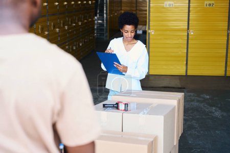 Téléchargez les photos : Gestionnaire afro-américain fait un inventaire des boîtes en carton en cours de dépôt, à côté d'un homme avec un chariot - en image libre de droit