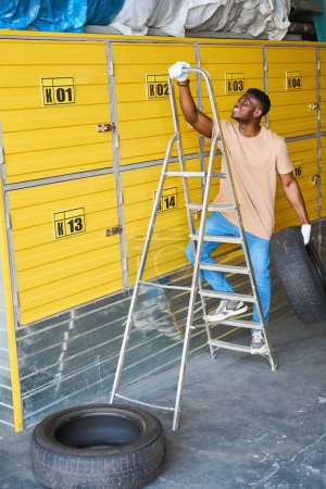 Téléchargez les photos : Homme afro-américain grimpant un escabeau avec un pneu de voiture à la main, des conteneurs dans un entrepôt jaune - en image libre de droit