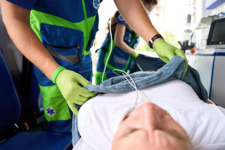 Photo for Medic covers a patient with a soft blanket in an ambulance, the patient lies on a special stretcher - Royalty Free Image