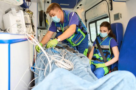Foto de El médico sujeta a un paciente en una ambulancia a una camilla con un cinturón de seguridad, una colega se sienta cerca - Imagen libre de derechos