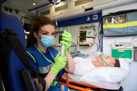 Foto de Empleado de un equipo de campo en una ambulancia prepara una jeringa con medicamentos, un paciente se acuesta con una máscara de oxígeno - Imagen libre de derechos