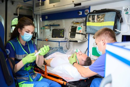 Foto de La doctora en una ambulancia prepara una jeringa con medicamentos, su colega sostiene una máscara de oxígeno en la cara de los pacientes - Imagen libre de derechos
