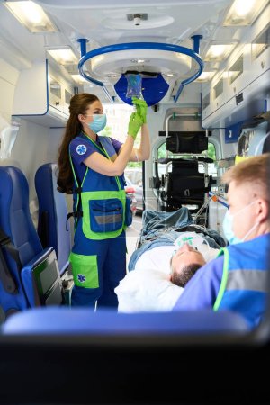 Téléchargez les photos : Une femme médecin met une intraveineuse sur une patiente dans une ambulance, à côté de son collègue dans un masque de protection - en image libre de droit