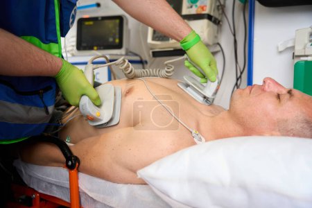Foto de Miembro del equipo de campo aplica electrodos de desfibrilador a un torso de pacientes mientras reanima al paciente en una ambulancia - Imagen libre de derechos