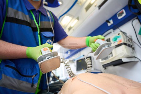 Téléchargez les photos : Homme ambulancier avec électrodes de défibrillateur dans ses mains, il effectue la réanimation d'un patient dans une ambulance - en image libre de droit