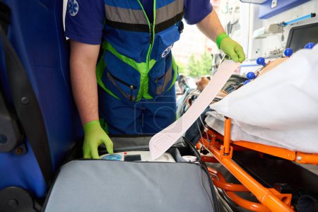 Foto de Un hombre con uniforme médico le hace un cardiograma a un hombre en una ambulancia, el paciente yace bajo un goteo - Imagen libre de derechos