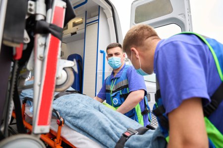 Téléchargez les photos : Médecins en uniforme déchargent une civière avec un patient d'une ambulance, il y a de l'équipement moderne dans la voiture - en image libre de droit