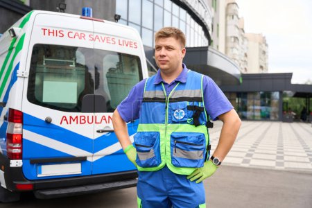 Photo for Young doctor of a visiting team stands by an ambulance, a man in a medical uniform and protective gloves - Royalty Free Image
