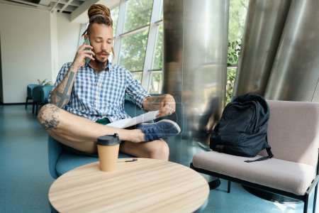 Photo for Guy is informal studying working documents and talking on phone in a coworking space, the room is spacious and bright - Royalty Free Image