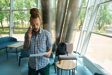 Photo for Man with dreadlocks studies work documents and communicates on the phone, he is in a comfortable coworking area - Royalty Free Image