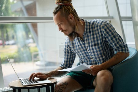 Photo for Guy in a plaid shirt works on a laptop in the coworking area, he is located at the viewport - Royalty Free Image