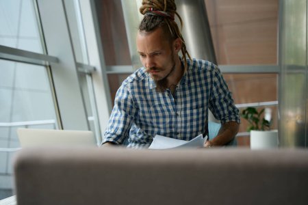 Téléchargez les photos : Homme en chemise à carreaux travaille sur un ordinateur portable dans la zone de coworking, il est situé à la fenêtre - en image libre de droit