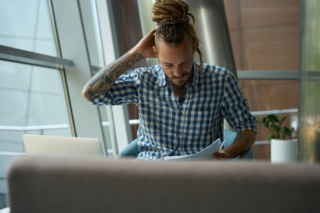 Téléchargez les photos : Freelance dans une chemise à carreaux travaille sur un ordinateur portable dans la zone de coworking, il est situé à la fenêtre - en image libre de droit