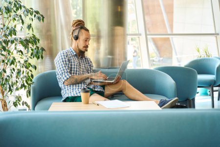 Photo for Informal IT specialist settled down with work on a cozy sofa, he uses a modern laptop and headset - Royalty Free Image