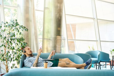 Photo for Young freelancer works lying on a couch in a coworking space, he uses a modern laptop and headset - Royalty Free Image