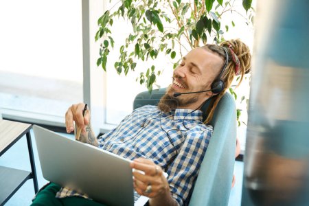 Photo for Freelancer with dreadlocks works lying on a couch in a coworking space, he uses a modern laptop and headset - Royalty Free Image