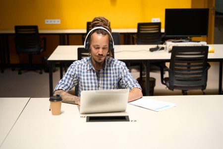 Foto de El hombre informal con rastas en la cabeza se estableció con el portátil en el escritorio de la oficina, el tipo utiliza un auricular para el trabajo - Imagen libre de derechos
