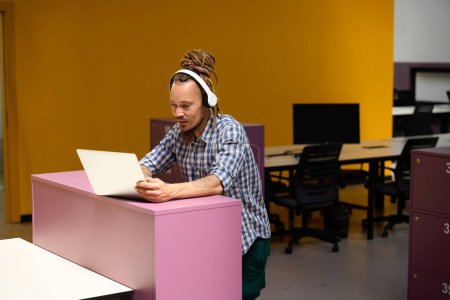 Téléchargez les photos : Le gars avec dreadlocks communique en ligne dans un bureau rose, il utilise un casque pour le travail - en image libre de droit