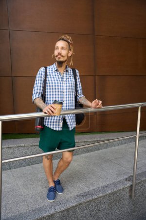 Foto de Freelancer se para con un vaso de café fuera de un edificio moderno, tiene una mochila y un tubo para dibujos - Imagen libre de derechos