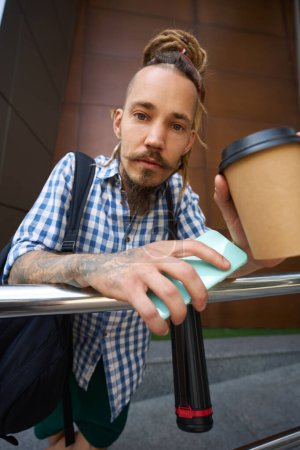 Foto de El hombre con rastas está fuera de un edificio moderno, tiene un vaso de café y un teléfono - Imagen libre de derechos