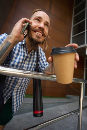 Photo for Guy with dreadlocks is on phone outside modern building, he has a glass of coffee and a tube for blueprints - Royalty Free Image