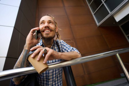 Photo for Guy in plaid shirt is onphone outside modern building, he has a glass of coffee and a tube for blueprints - Royalty Free Image