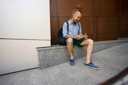 Foto de Sonriendo freelancer charlando en línea por teléfono en una acera de granito cerca de un edificio moderno - Imagen libre de derechos