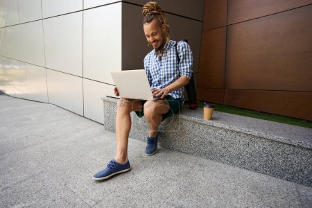 Foto de Feliz freelancer charlando en línea en una acera de granito cerca de un edificio moderno, junto a un vaso de café - Imagen libre de derechos