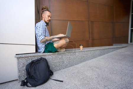 Photo for Informal guy works on laptop on granite curb near modern building, next to a backpack and a glass of coffee - Royalty Free Image