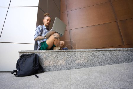 Foto de Hombre en una camisa a cuadros chats en línea en una acera de granito cerca de un edificio moderno - Imagen libre de derechos