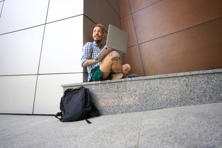 Foto de Chico en una camisa a cuadros chats en línea en una acera de granito cerca de un edificio moderno - Imagen libre de derechos